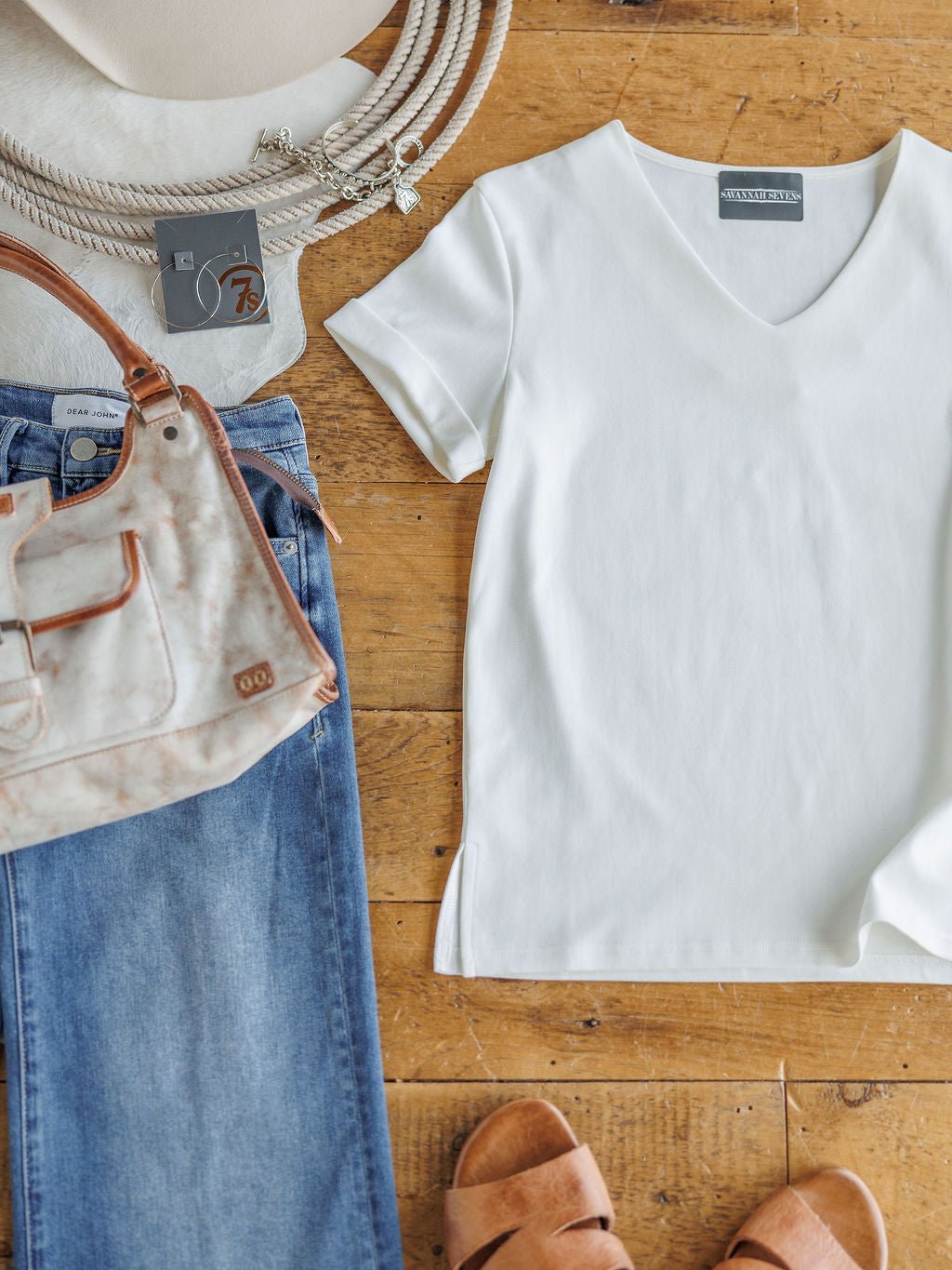 A flat lay featuring The Kinston by Savannah Sevens western life{&}style: an off-white short sleeve top, blue jeans, tan sandals, beige handbag and hat, plus a beaded necklace on a wooden floor.