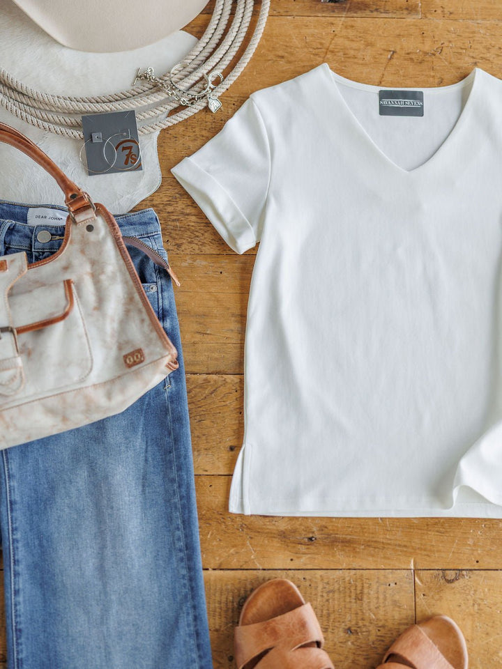 A flat lay featuring The Kinston by Savannah Sevens western life{&}style: an off-white short sleeve top, blue jeans, tan sandals, beige handbag and hat, plus a beaded necklace on a wooden floor.