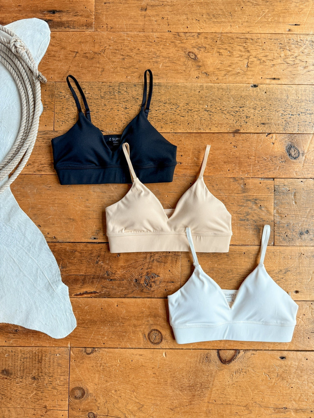 The Kirby bralettes by Savannah Sevens western life{&}style in black, beige, and white, featuring wide elastic bands and removable padding, are displayed on a wooden floor beside a coiled rope and white textured cloth.