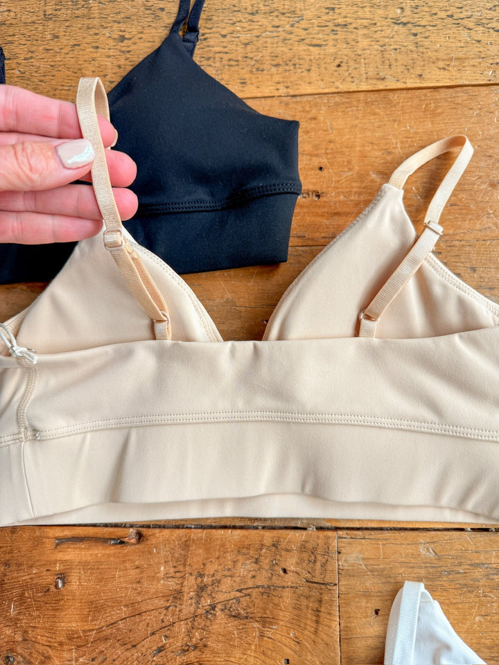 A hand adjusts the strap of The Kirby beige bralette by Savannah Sevens western life{&}style, featuring a wide elastic band and displayed on wood, with a black bralette and part of a white garment partially visible nearby.