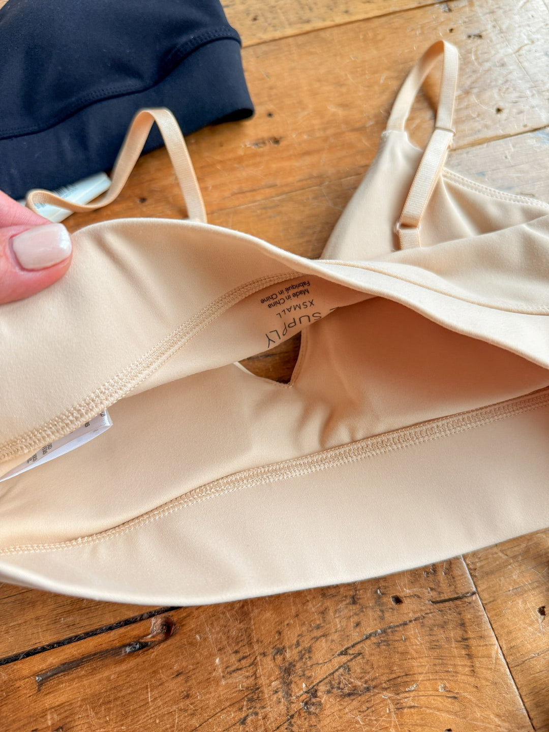 A close-up of The Kirby by Savannah Sevens western life{&}style, a beige stretch t-shirt bra with an inner pocket and small opening, lies on wood. A hand holds one edge; part of a black bra appears in the background.