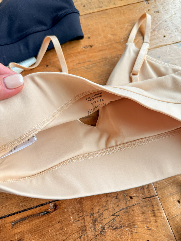 A close-up of The Kirby by Savannah Sevens western life{&}style, a beige stretch t-shirt bra with an inner pocket and small opening, lies on wood. A hand holds one edge; part of a black bra appears in the background.