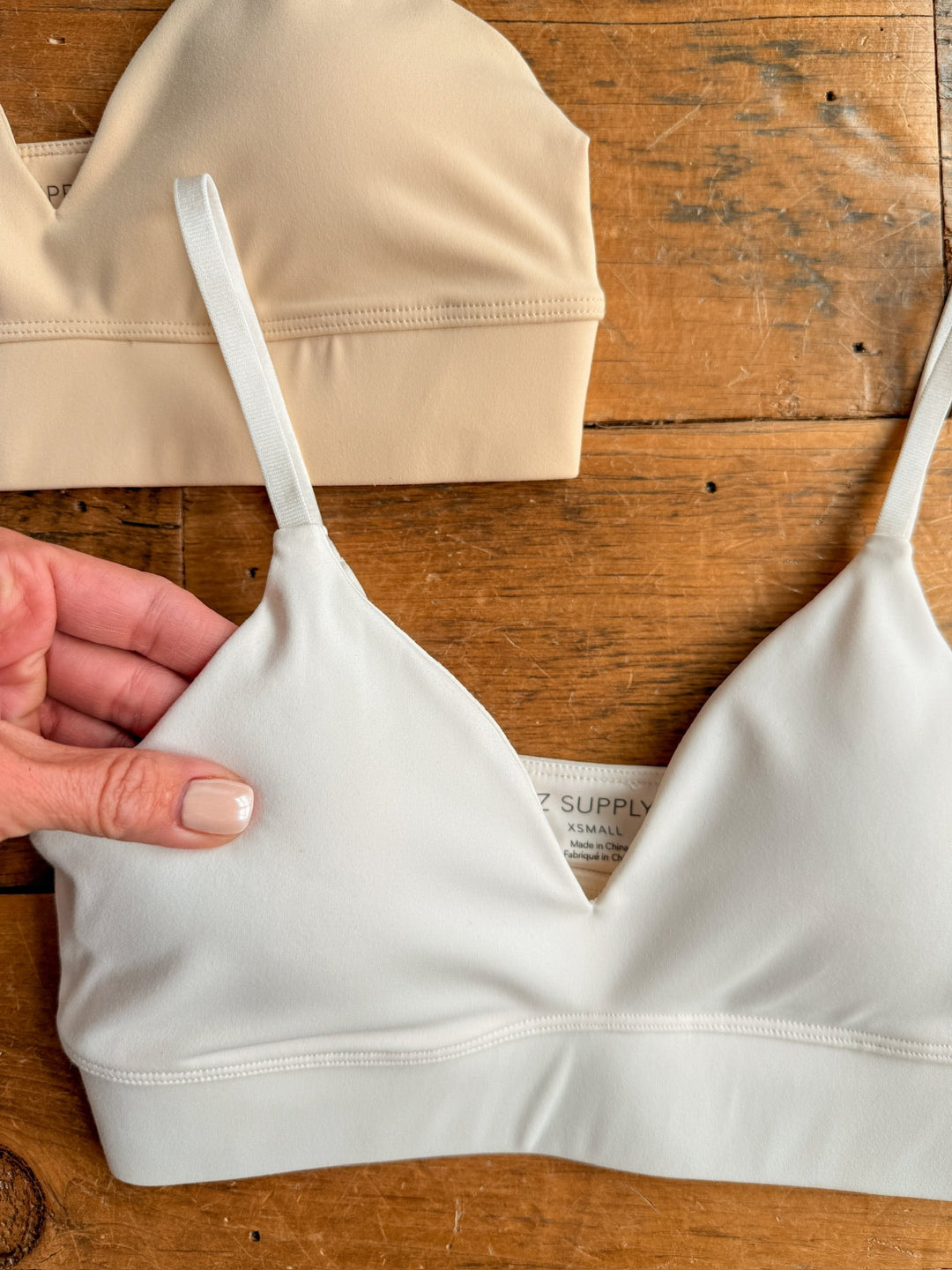 A hand displays The Kirby bralette in white from Savannah Sevens western life{&}style with a wide elastic band on wood; above it lies the beige version. Both have thin straps and a soft, seamless design.