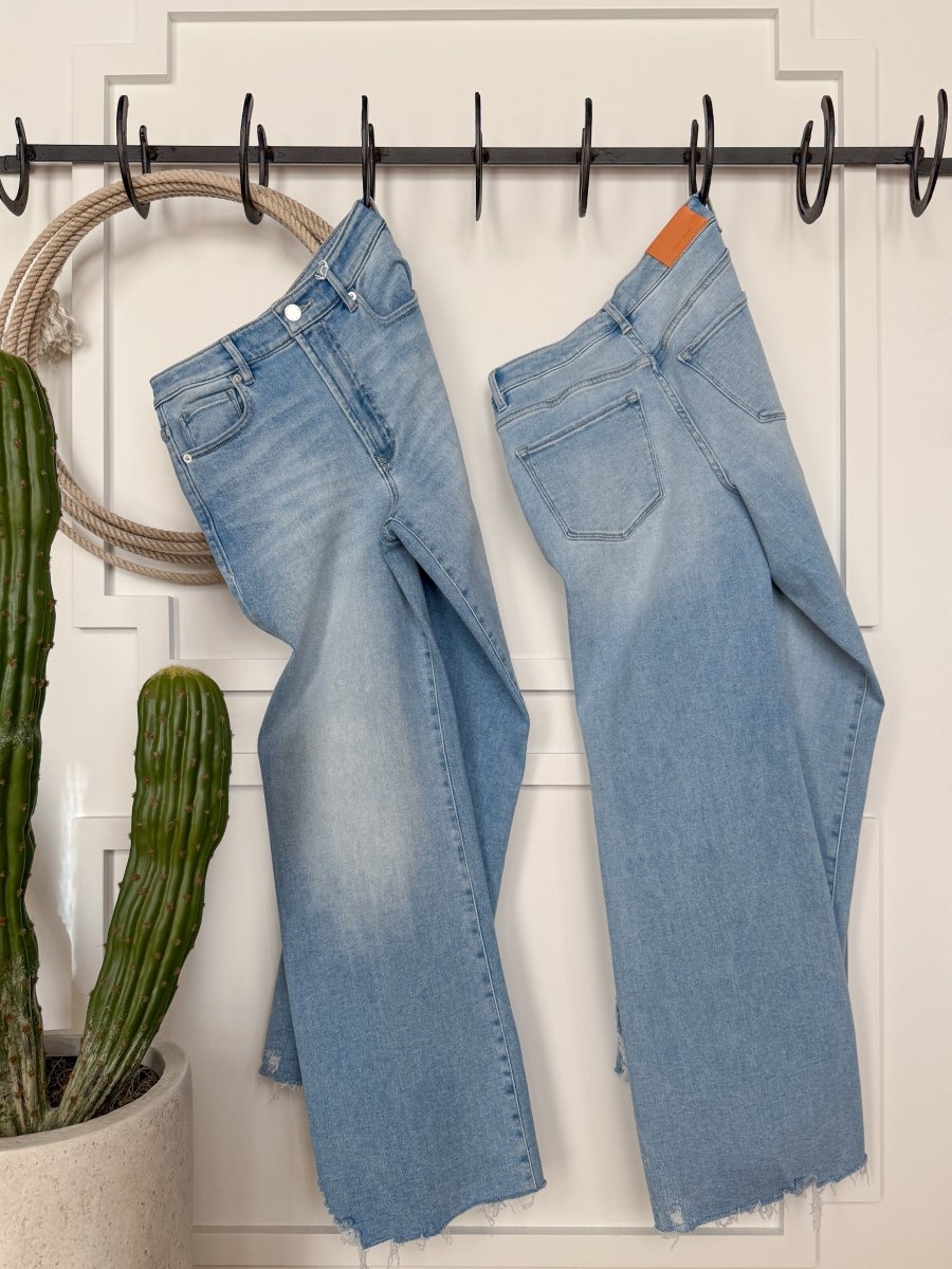 Two pairs of The Kollins High Rise Wide Leg Jeans by Savannah Sevens western life{&}style, in light blue with frayed hems, hang by their belt loops on black hooks against a white paneled wall. A potted cactus and coiled rope sit to the left.