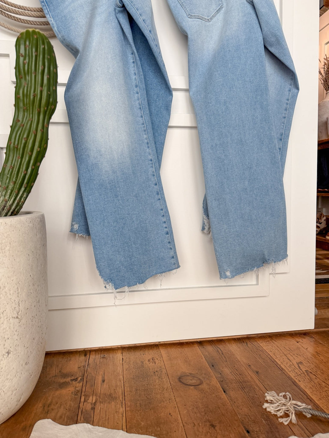 The Kollins High Rise Wide Leg Jean by Savannah Sevens western life{&}style in light blue denim with frayed hems hangs on a white paneled wall above a wooden floor, next to a tall green cactus in a white pot and knotted rope in the corner.