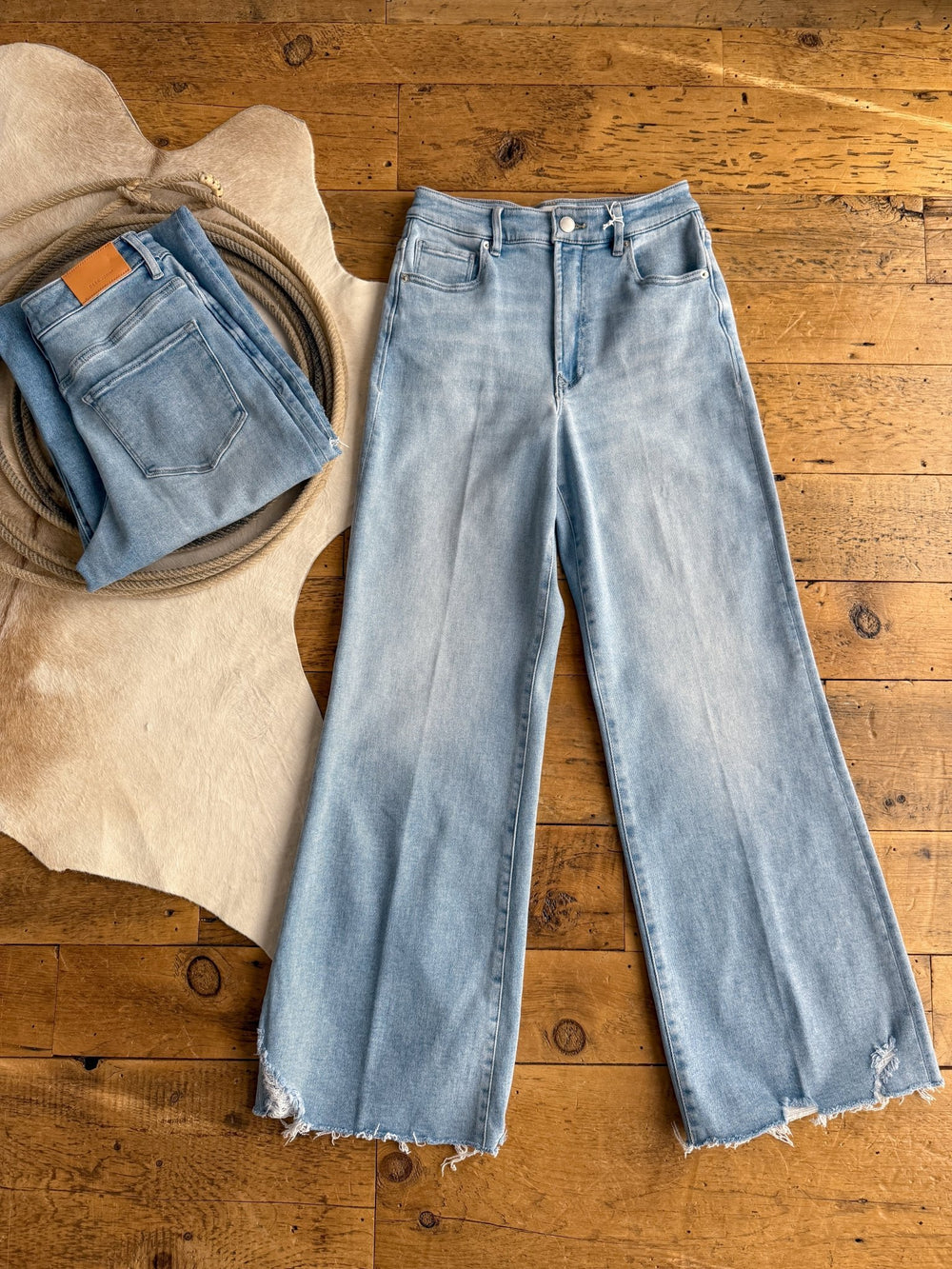The Kollins High Rise Wide Leg Jean by Savannah Sevens western life{&}style is laid flat with frayed hems on a wooden floor, beside a folded pair of high rise denim jeans on a textured mat and tan leather-like rug.