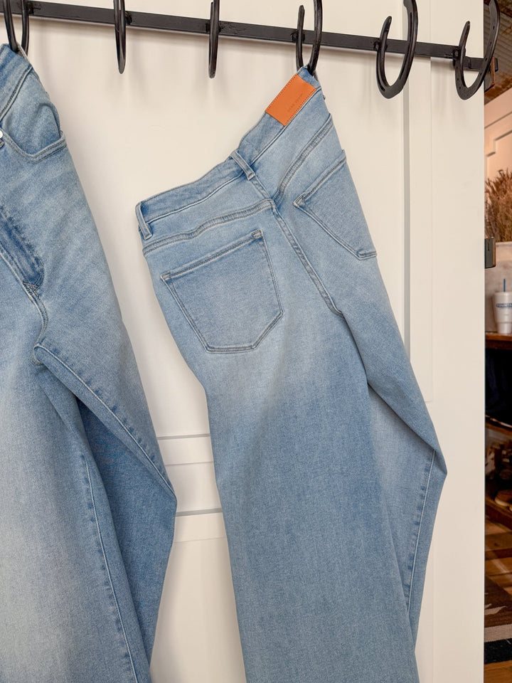 The Kollins High Rise Wide Leg Jean by Savannah Sevens western life{&}style in light blue hangs by the belt loops on black hooks against a white door, with part of another wide leg jean visible on the left.