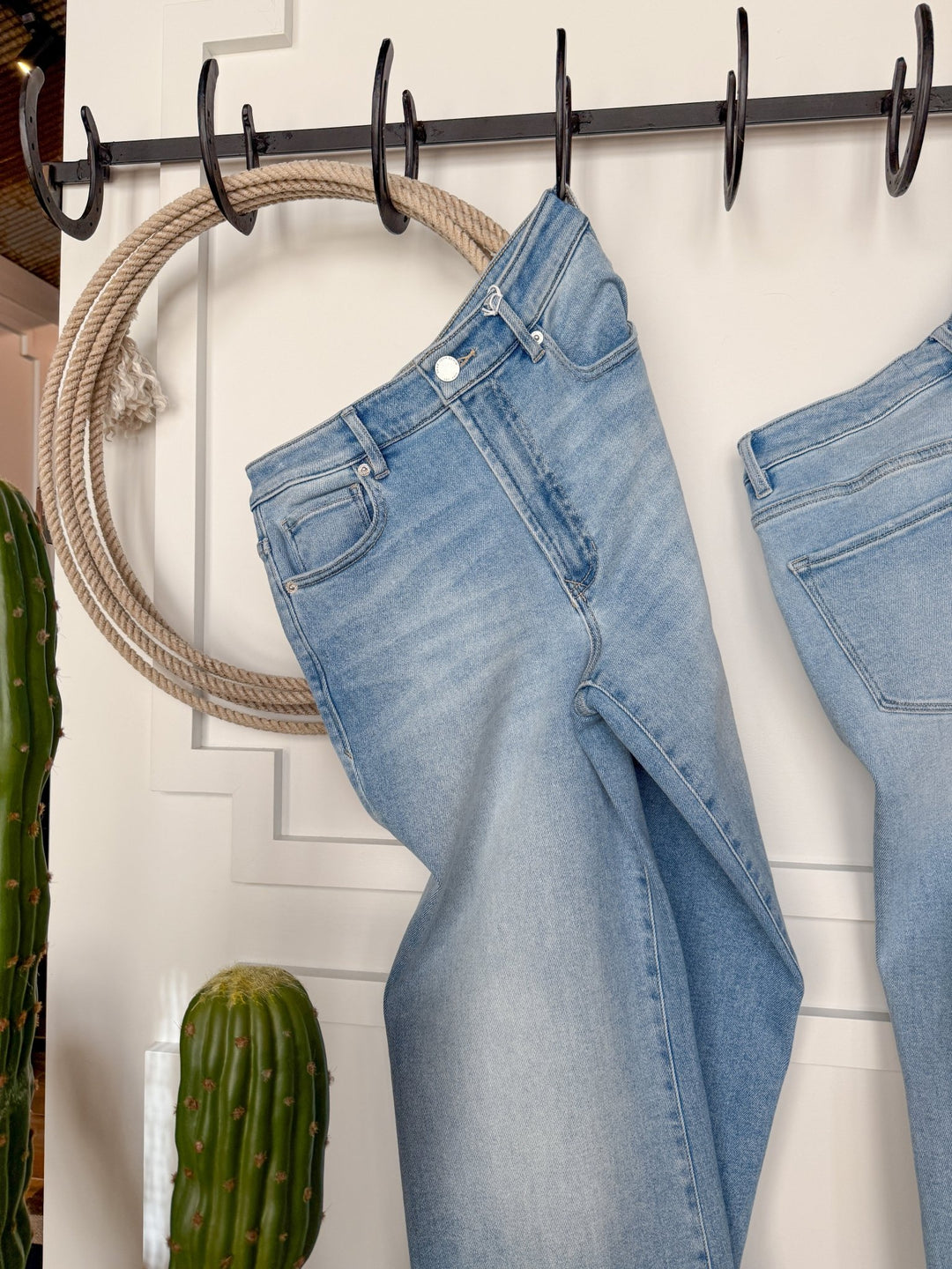 The Kollins High Rise Wide Leg Jean by Savannah Sevens western life{&}style in light blue hangs on a black hook near a coiled rope and two green cacti, set against a white wall with decorative molding.