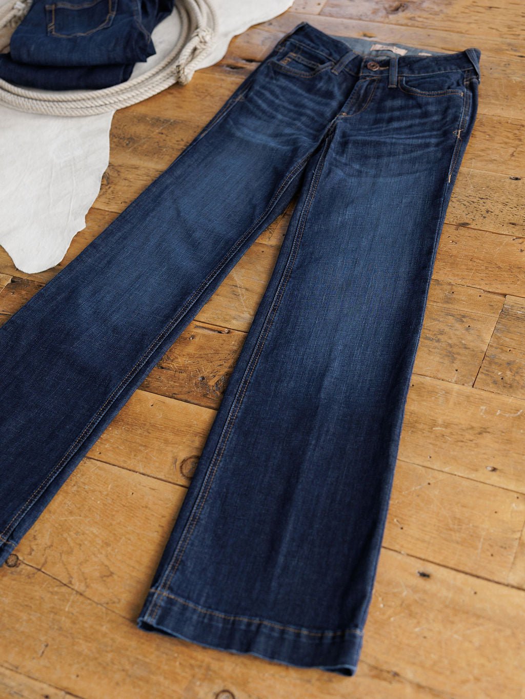 A pair of Ariat The Kolt Perfect Rise Trouser Jeans in a medium dark wash lies flat on a wooden floor, with another folded pair and a coiled rope visible in the background.