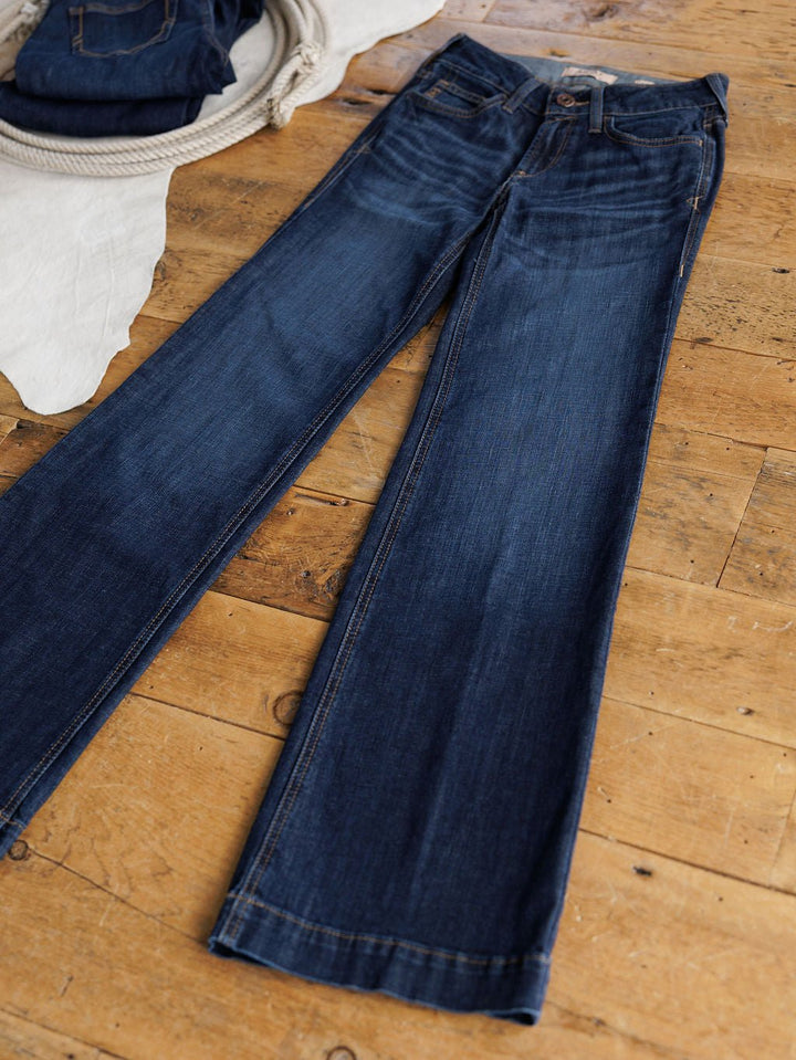 A pair of Ariat The Kolt Perfect Rise Trouser Jeans in a medium dark wash lies flat on a wooden floor, with another folded pair and a coiled rope visible in the background.