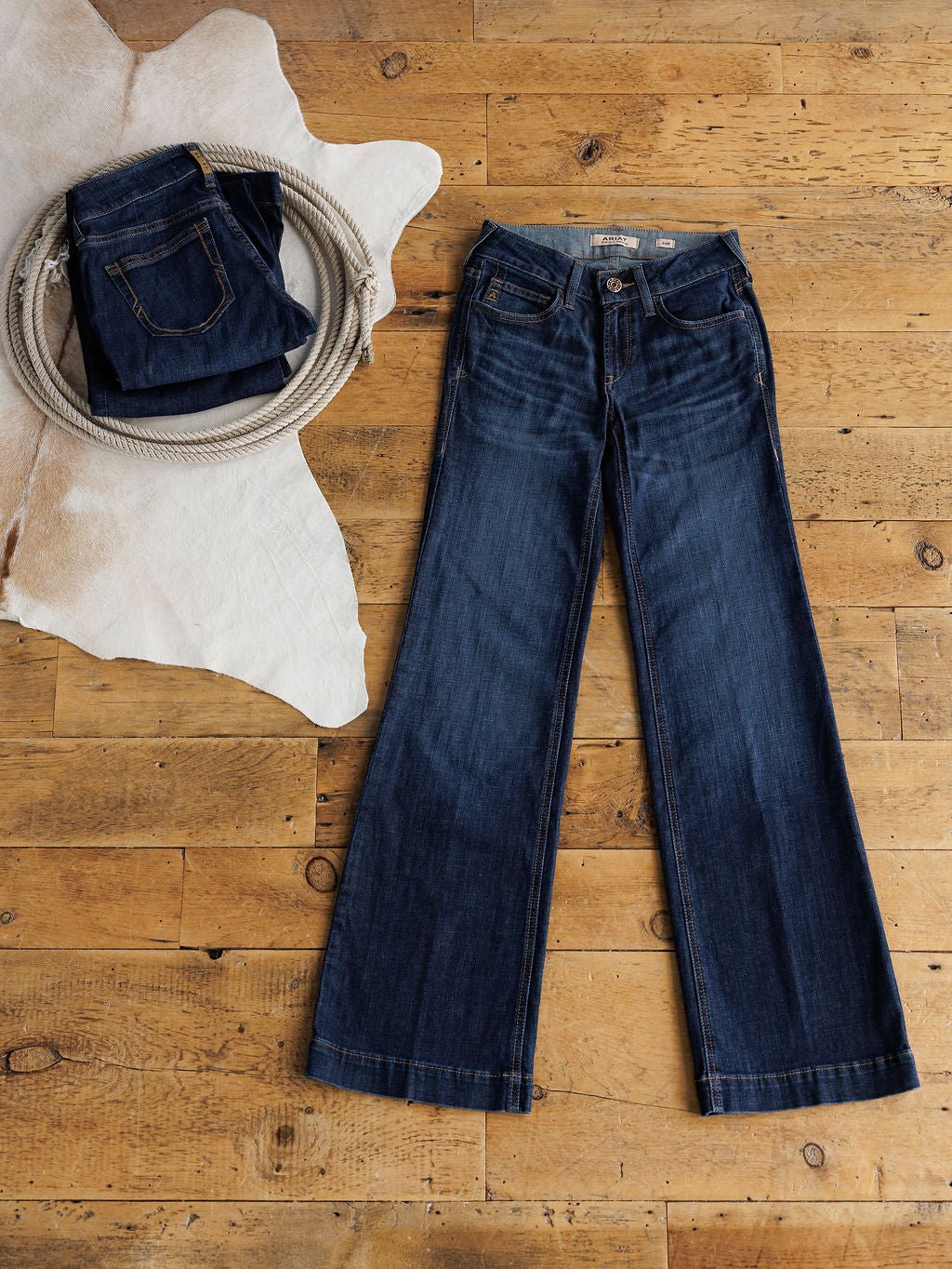 A pair of medium dark wash jeans lies flat on a wooden floor. Nearby, Ariat’s The Kolt Perfect Rise Trouser Jean sits folded on a white cowhide rug, partially wrapped with a coiled rope.