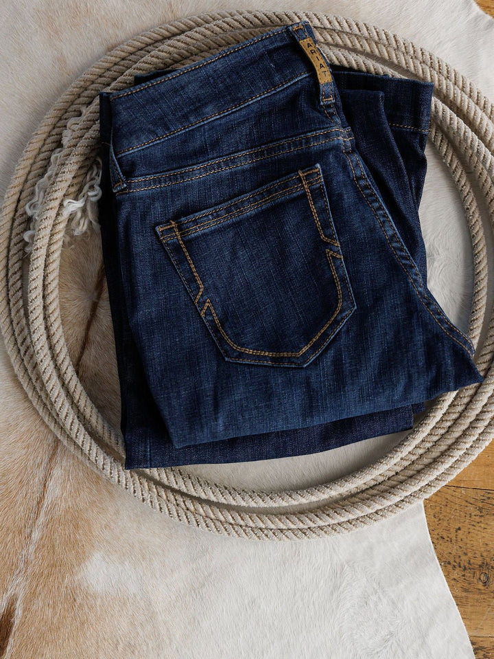 A pair of Ariat The Kolt Perfect Rise Trouser Jeans in medium dark wash with tan stitching is folded on a coiled rope, displayed on a light brown and white cowhide rug.