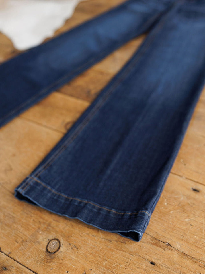 The Ariat Kolt Perfect Rise Trouser Jean, in a medium dark wash with flared legs, is laid flat on a rustic wooden floor, showcasing visible seam details and a slightly frayed hem.