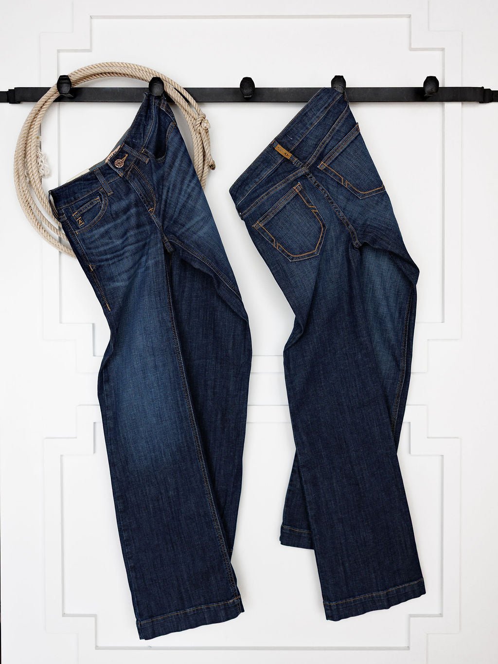Two pairs of blue jeans, including the Ariat Kolt Perfect Rise Trouser Jean in a medium dark wash, hang from a black rod on a white wall, with a coiled rope draped over the left side.