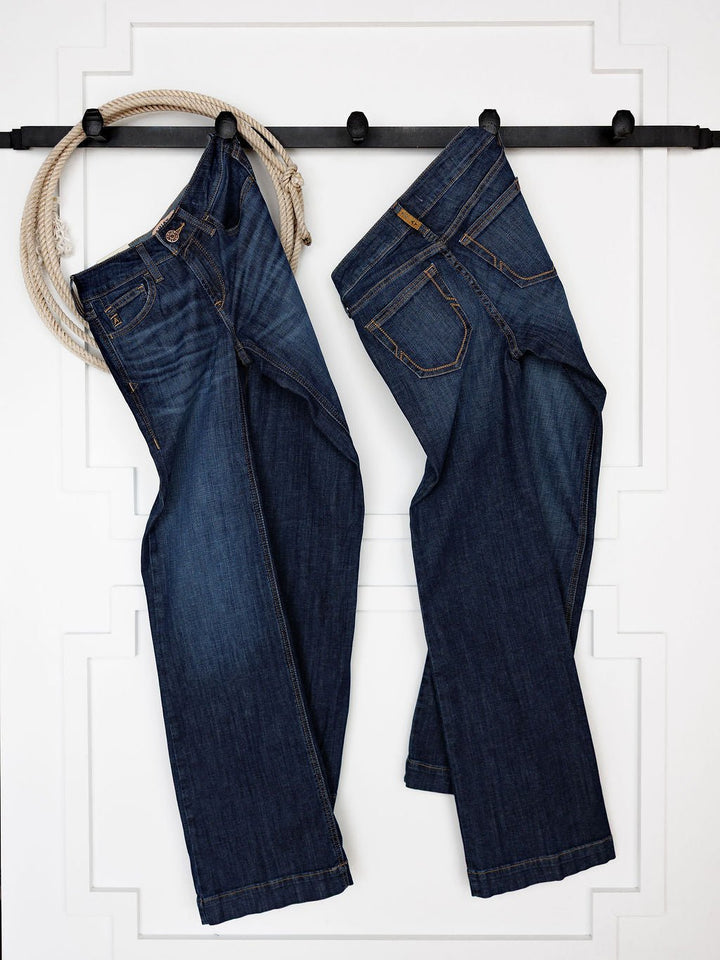 Two pairs of blue jeans, including the Ariat Kolt Perfect Rise Trouser Jean in a medium dark wash, hang from a black rod on a white wall, with a coiled rope draped over the left side.