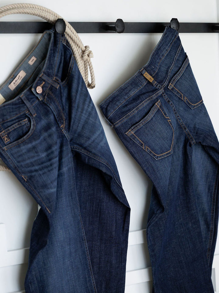 Two pairs of Ariat The Kolt Perfect Rise Trouser Jeans in a medium dark wash hang side by side on black hooks against a white wall, with a coiled beige rope beside them. These jeans offer the ideal rise for everyday comfort.