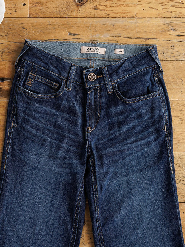 A pair of Ariat The Kolt Perfect Rise Trouser Jeans in medium dark wash with tan stitching and button fly, marked size 24R, is displayed flat on a wooden surface with an ARIAT label inside the waistband.