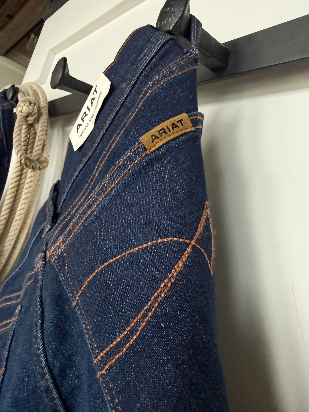 Close-up of dark blue Kyndal Perfect Rise Trouser Jeans by Savannah Sevens western life{&}style hanging on a black hook, showcasing brown stitching details and two visible brand labels. Made from durable UltraStretch fabric, these jeans are displayed alongside a coiled rope hanging on the left side of the image.
