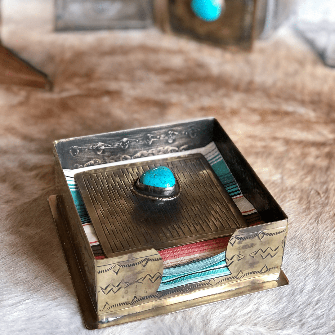 The La Puebla Cocktail Napkin Holder by Savannah Sevens western life{&}style features engraved German silver with a turquoise stone, holding colorful striped napkins on tan fur and highlighting classic southwest style.