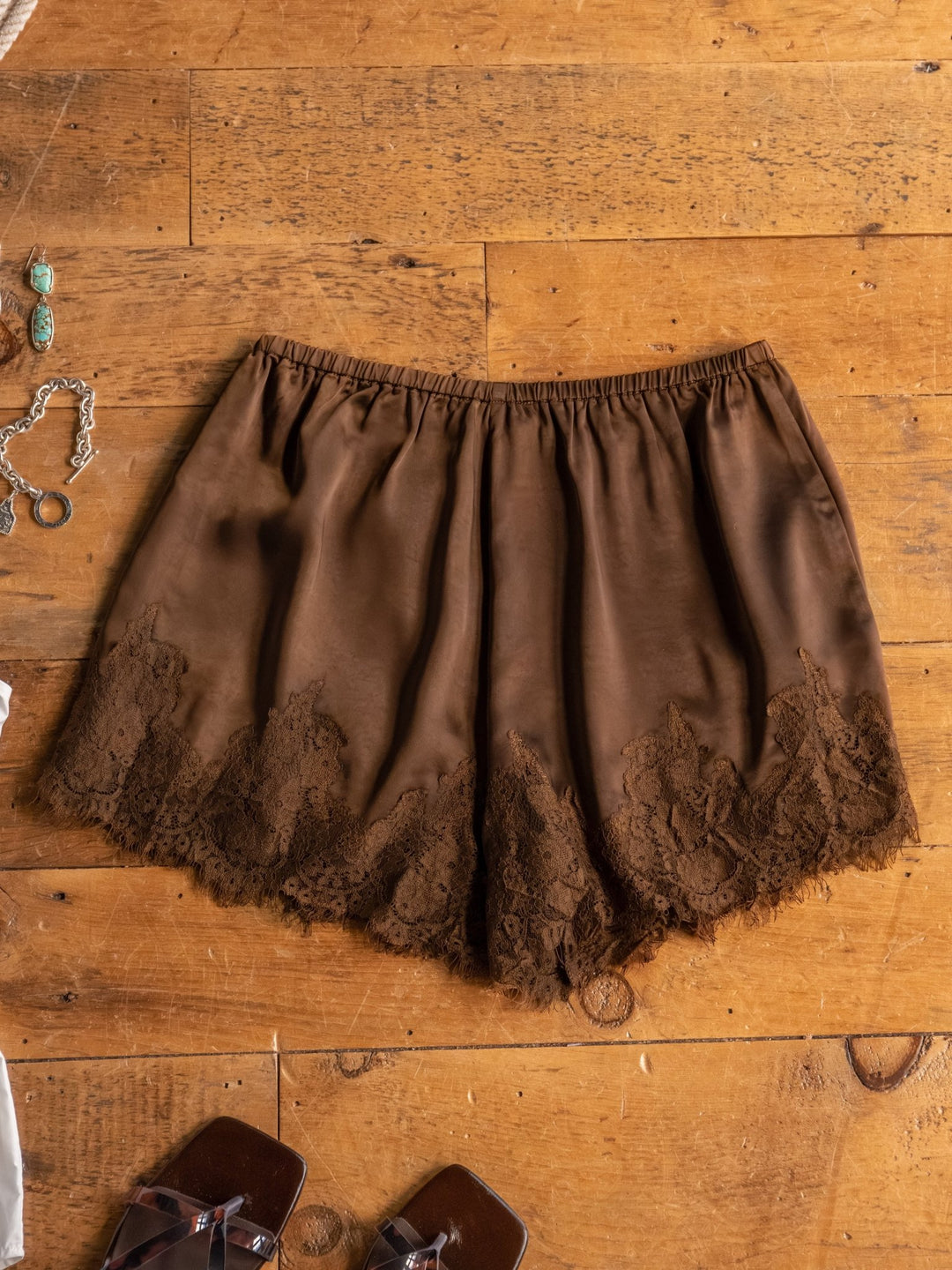 The Laconia satin shorts by Savannah Sevens western life{&}style are laid flat on a wooden floor, detailed with a lace hem and styled with jewelry, a ring, and brown sandals at the bottom of the image.