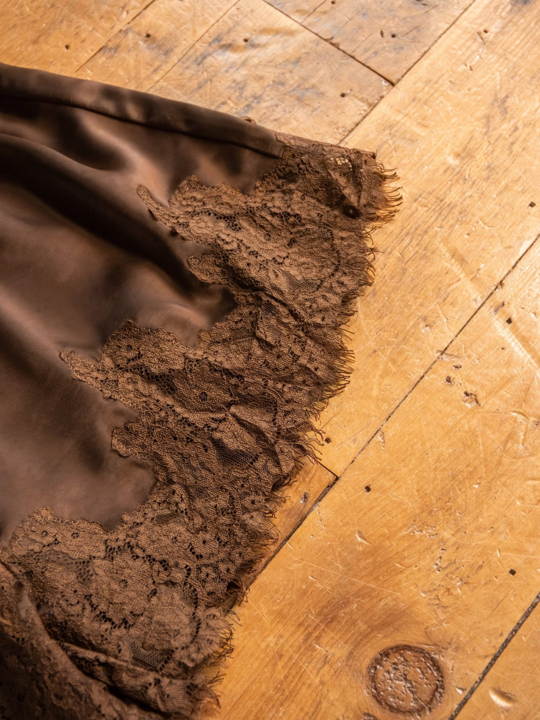 A close-up of The Laconia chocolate satin shorts by Savannah Sevens western life{&}style, featuring a delicate lace hem, draped over a wooden floor with visible grain and knots.