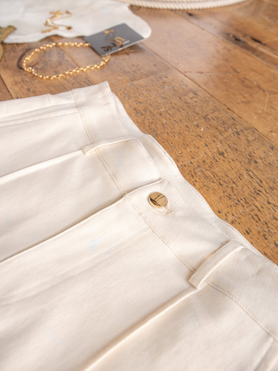 Close-up of The Ladora cream-colored pintuck shorts by Savannah Sevens western life{&}style, featuring a gold button on a wooden surface. Jewelry and accessories are slightly visible in the background.
