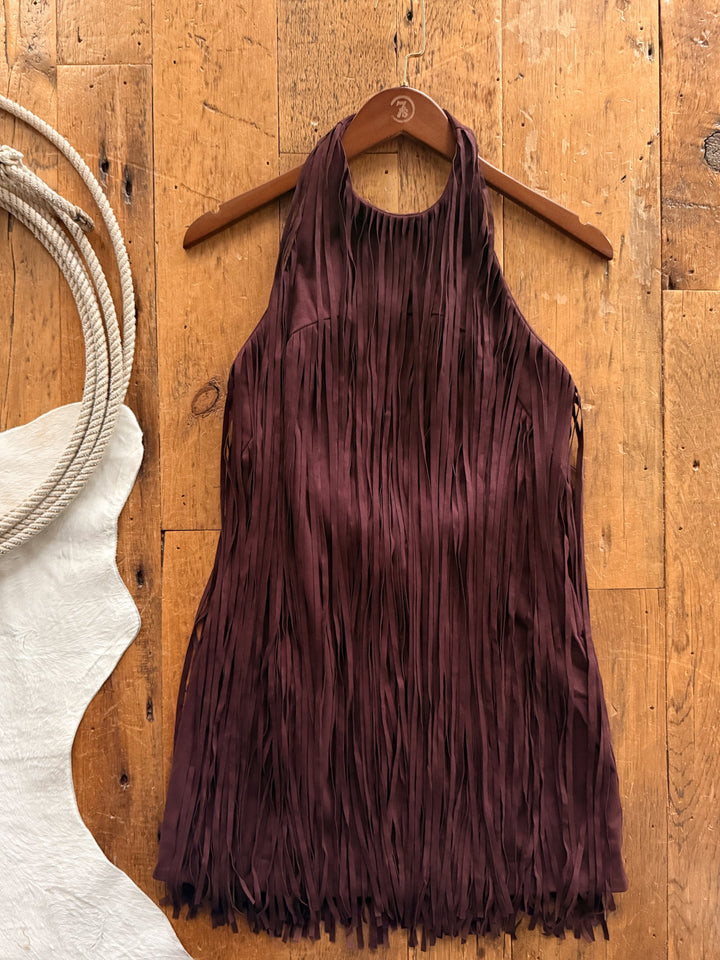 A Lahoma Black Cherry Fringe Dress hanging on a wooden hanger against a wooden floor with a coiled rope and a white hide.