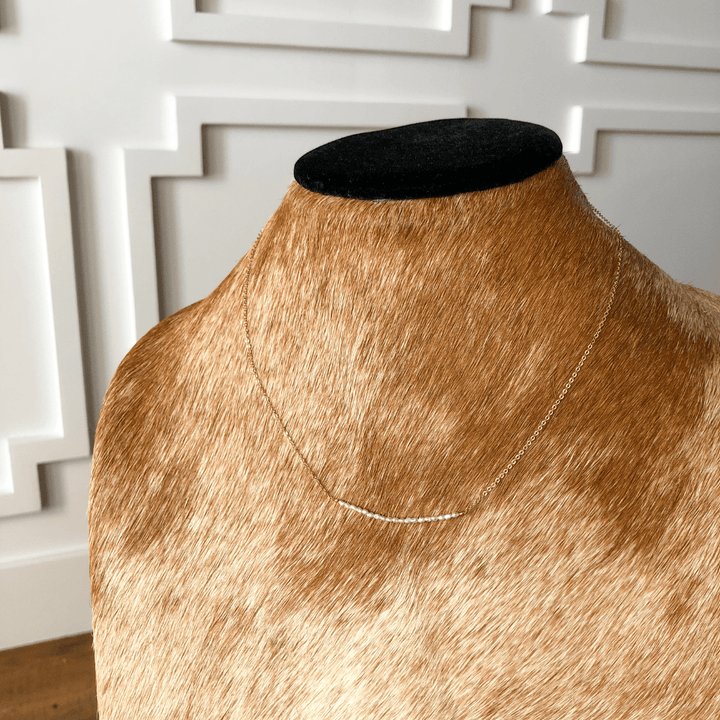 The Lahoma Necklace by Savannah Sevens western life{&}style, featuring a slim gold-filled horizontal bar pendant, is showcased on a brown cowhide bust against a white decorative wall.