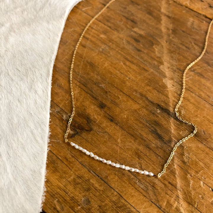 The Lahoma Necklace by Savannah Sevens western life{&}style, a delicate gold-filled piece with small white beads, is displayed on a rustic wooden surface next to soft textured fabric.