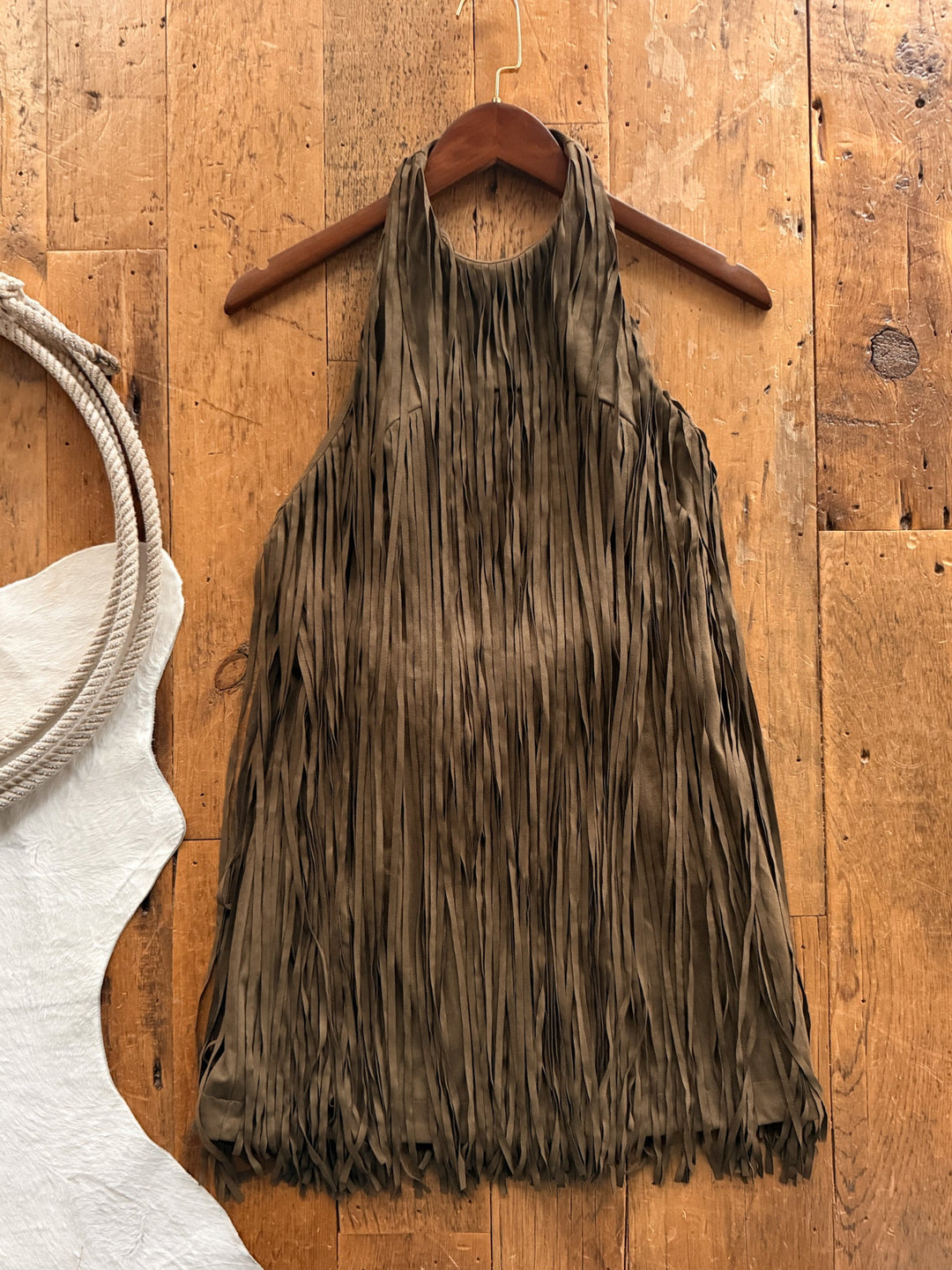 Lahoma Olive Fringe Dress in olive faux suede, a halter-neck style completely covered in fringe, hanging on a wooden hanger against a wood floor.