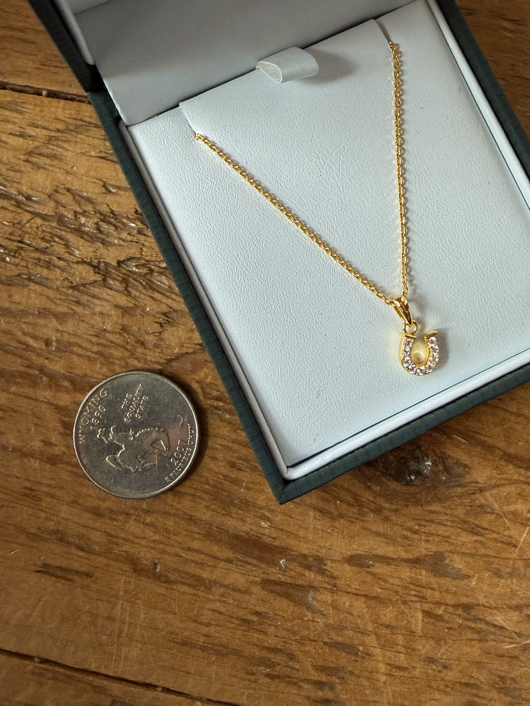 The Lajitas Gold Horseshoe Pendant Necklace by Savannah Sevens western life{&}style is displayed in a white jewelry box on a wooden surface, shown beside a 25-cent Canadian coin for size reference.