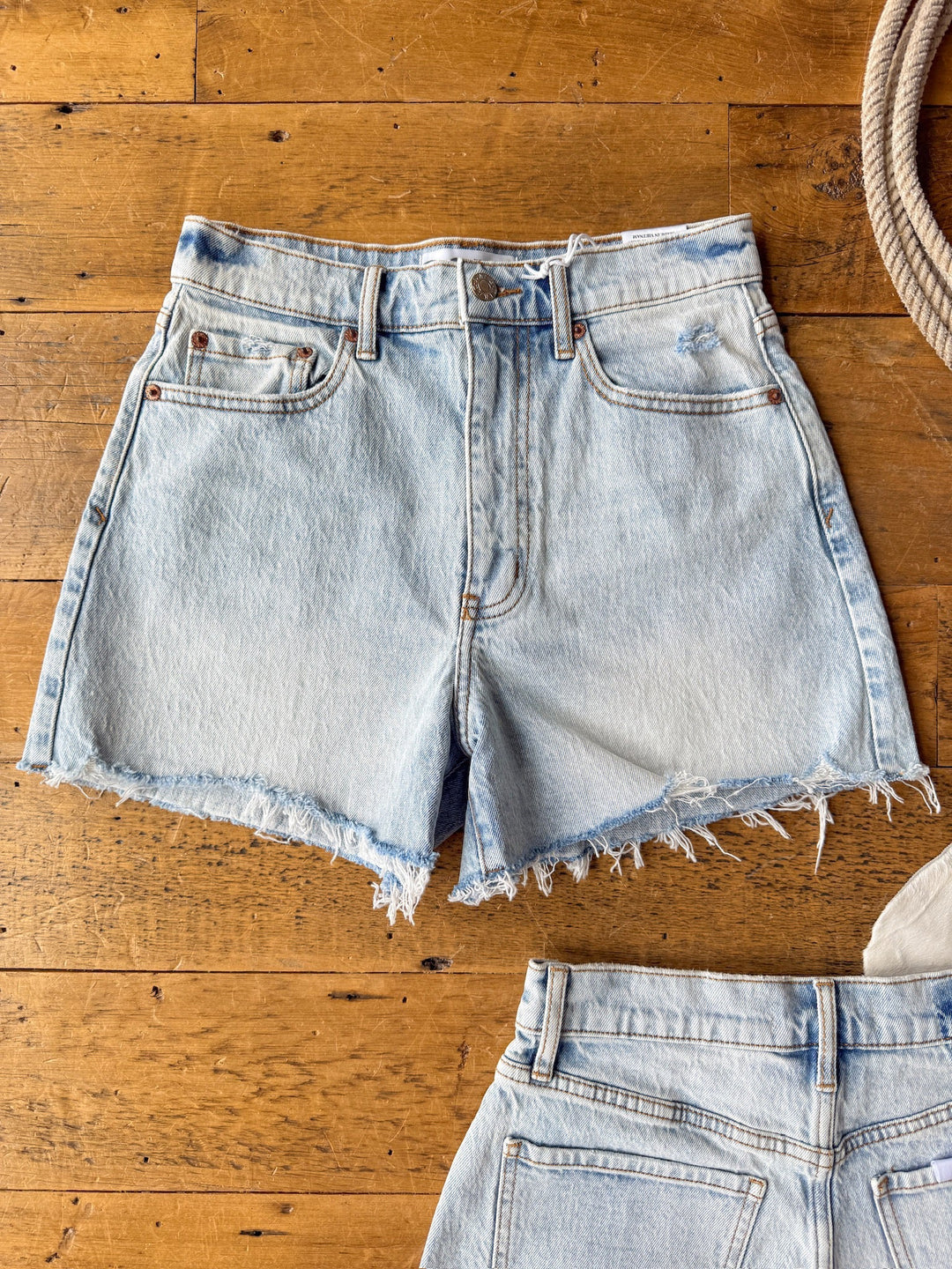 The Laken Shorts by Savannah Sevens western life{&}style—a pair of light wash denim shorts with raw hems—rest on a wooden surface, with another pair of shorts and a coil of rope partially visible nearby.