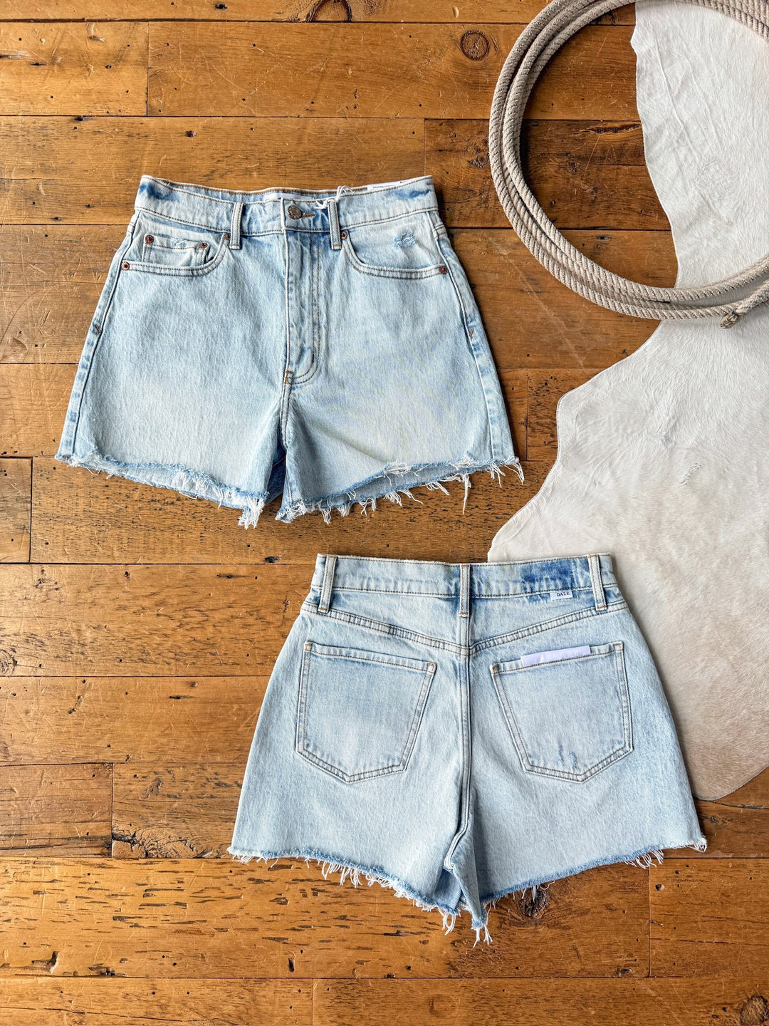 Two pairs of The Laken Shorts by Savannah Sevens western life{&}style, light wash denim with raw hems, are displayed front and back on a wooden floor, accompanied by a coiled rope and a light fabric.