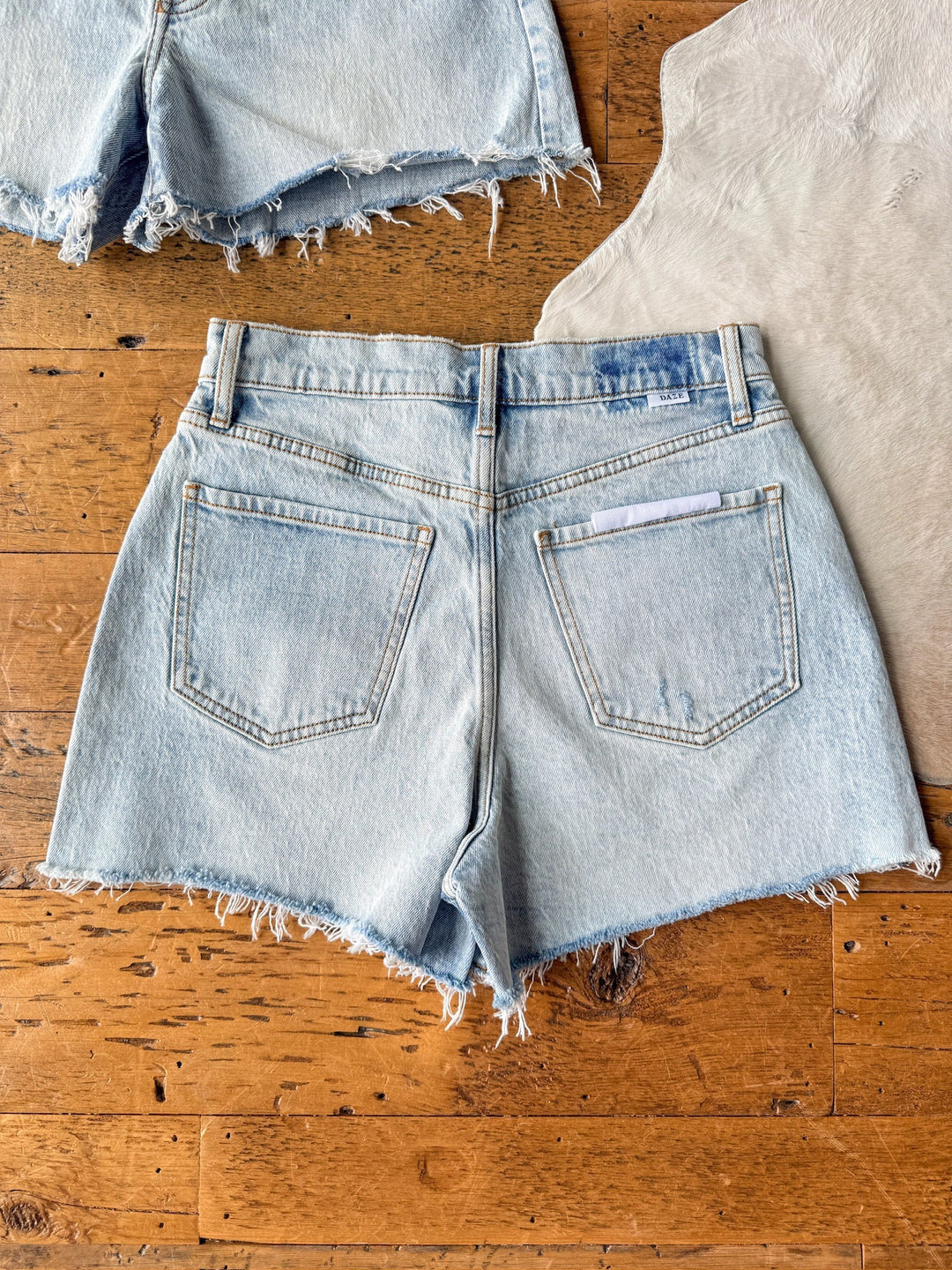 The Laken Shorts by Savannah Sevens western life{&}style are light wash, high rise denim shorts with raw hems and two back pockets, shown laid flat on a wooden floor beside another denim piece and a light-colored fabric.