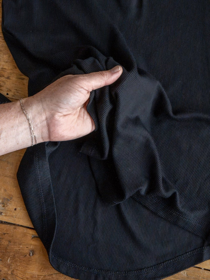 A hand wearing a thin bracelet grips a section of black ribbed fabric from the Lakota Black Ribbed Top by Savannah Sevens western life{&}style, which is spread out on a wooden surface.