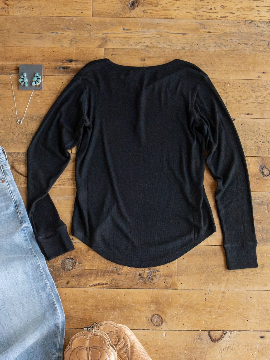 The Lakota Black Ribbed Top by Savannah Sevens western life{&}style is laid flat on a wooden floor, styled with a pair of jeans, a quilted tan purse, turquoise earrings, and a matching necklace placed nearby.