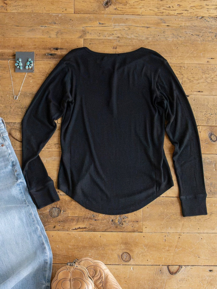 The Lakota Black Ribbed Top by Savannah Sevens western life{&}style is laid flat on a wooden floor, styled with a pair of jeans, a quilted tan purse, turquoise earrings, and a matching necklace placed nearby.