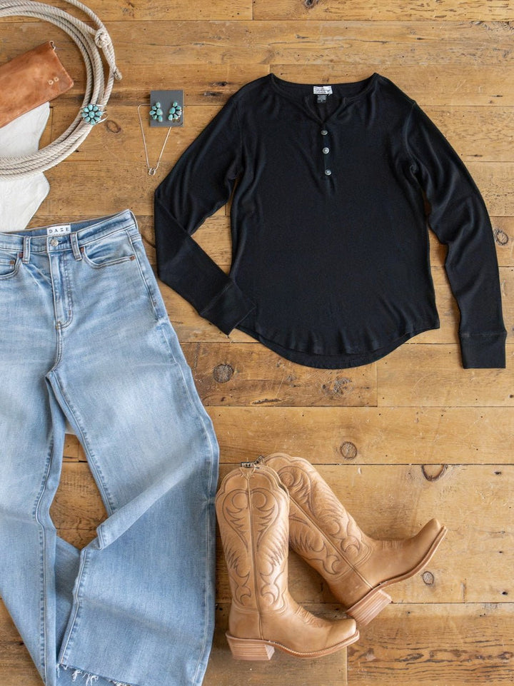 Flat lay featuring the Lakota Black Ribbed Top by Savannah Sevens western life{&}style, paired with light blue jeans, tan cowboy boots, turquoise jewelry, and a brown leather wallet on a wooden floor.