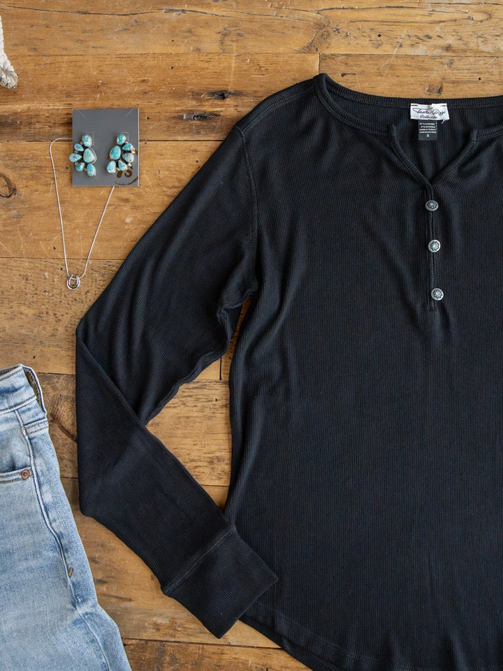 The Lakota Black Ribbed Top by Savannah Sevens western life{&}style, paired with blue jeans, turquoise earrings, and a silver necklace, is arranged on a rustic wooden surface.