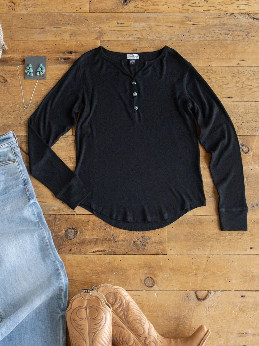 The Lakota Black Ribbed Top by Savannah Sevens western life{&}style is laid flat on a wooden floor alongside light blue jeans, tan cowboy boots, a necklace, and turquoise earrings. The ribbed long sleeve design adds texture to this classic western look.
