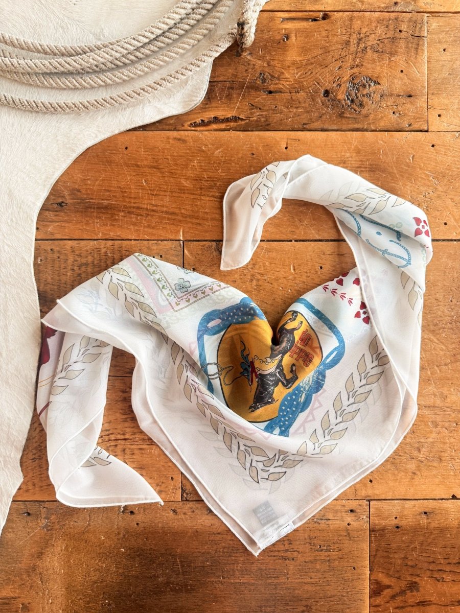 A Lamesa Neck Scarf by Savannah Sevens western life{&}style, featuring a patterned white sheer design with colorful western graphics, is shaped into a heart on wood beside a coiled rope and textured white fabric.