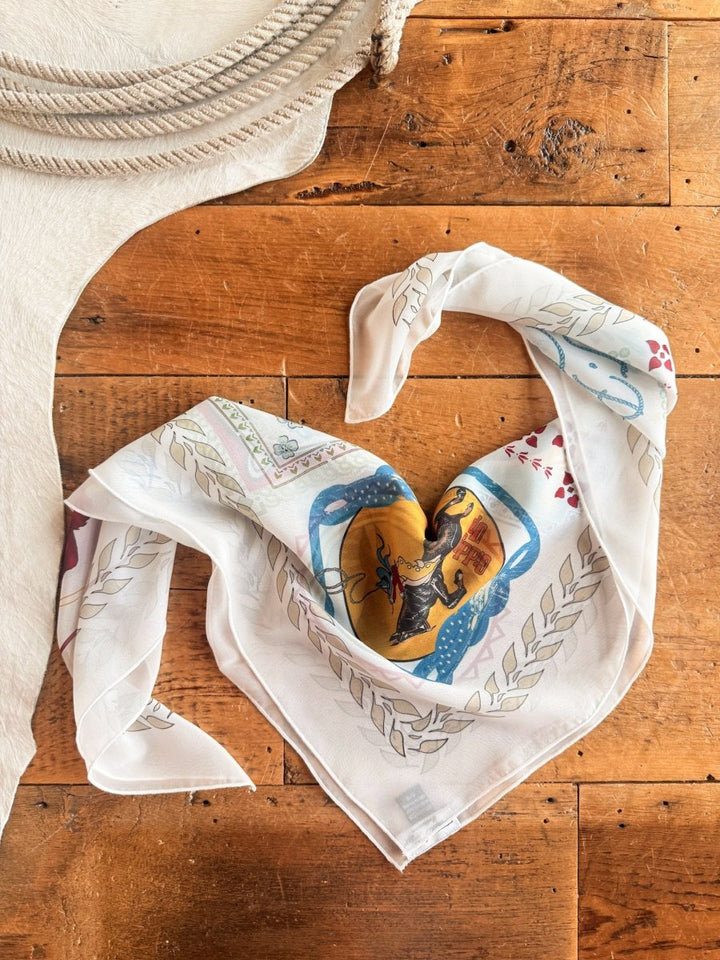 A Lamesa Neck Scarf by Savannah Sevens western life{&}style, featuring a patterned white sheer design with colorful western graphics, is shaped into a heart on wood beside a coiled rope and textured white fabric.