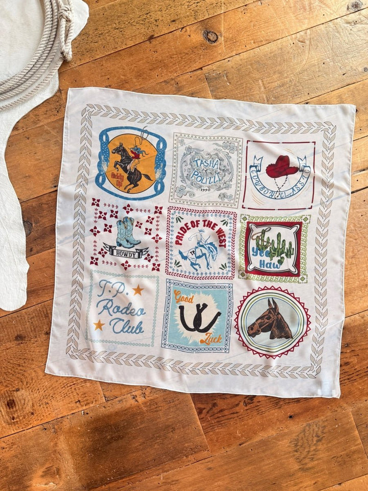 The Lamesa Neck Scarf by Savannah Sevens western life{&}style is a square scarf with nine graphic patches—cowboy, horses, cacti, red hat, and phrases like “Howdy” and “Good Luck”—displayed on a wooden floor.