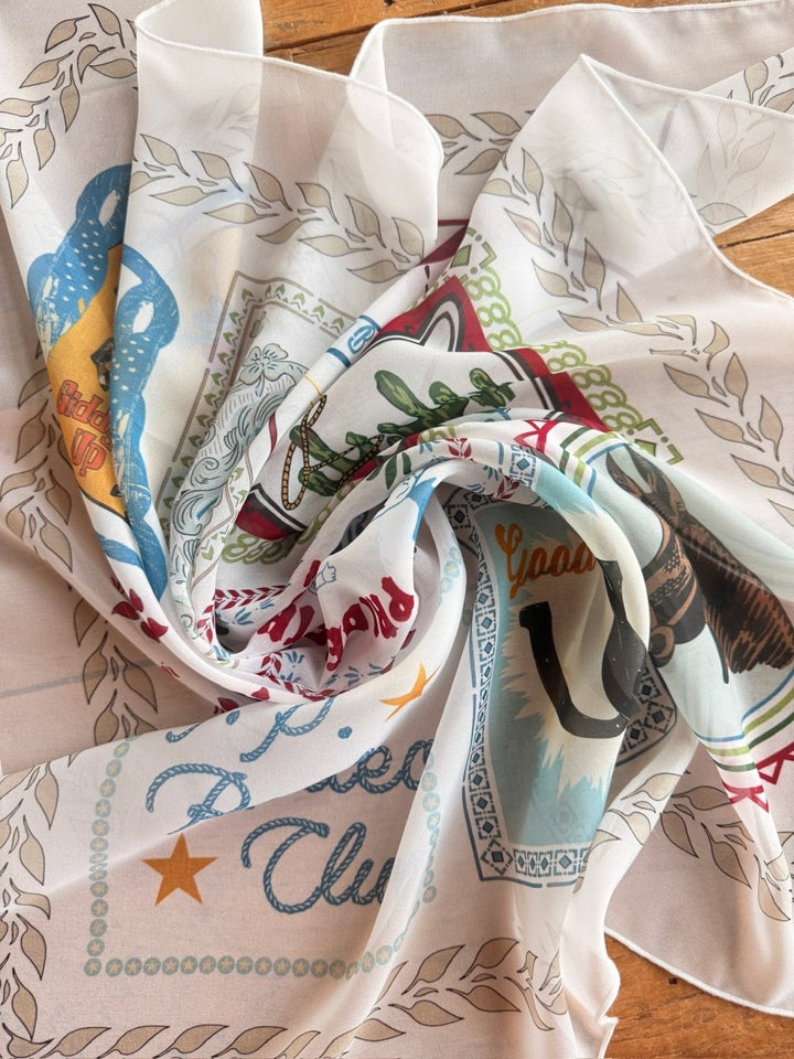 The Lamesa Neck Scarf by Savannah Sevens western life{&}style, with a colorful vintage-style western design—featuring a horse, cowboy hat, and stars—lies artfully crumpled on a wooden surface.
