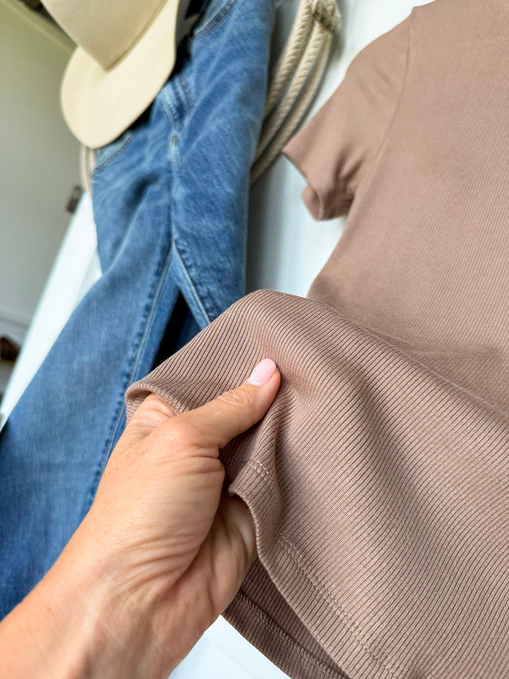 A hand holds the hem of the Lamotte Beige Ribbed Tee by Savannah Sevens western life{&}style, a short sleeve ribbed top with a built-in bra in beige, laid out alongside blue jeans and a beige hat in the background.