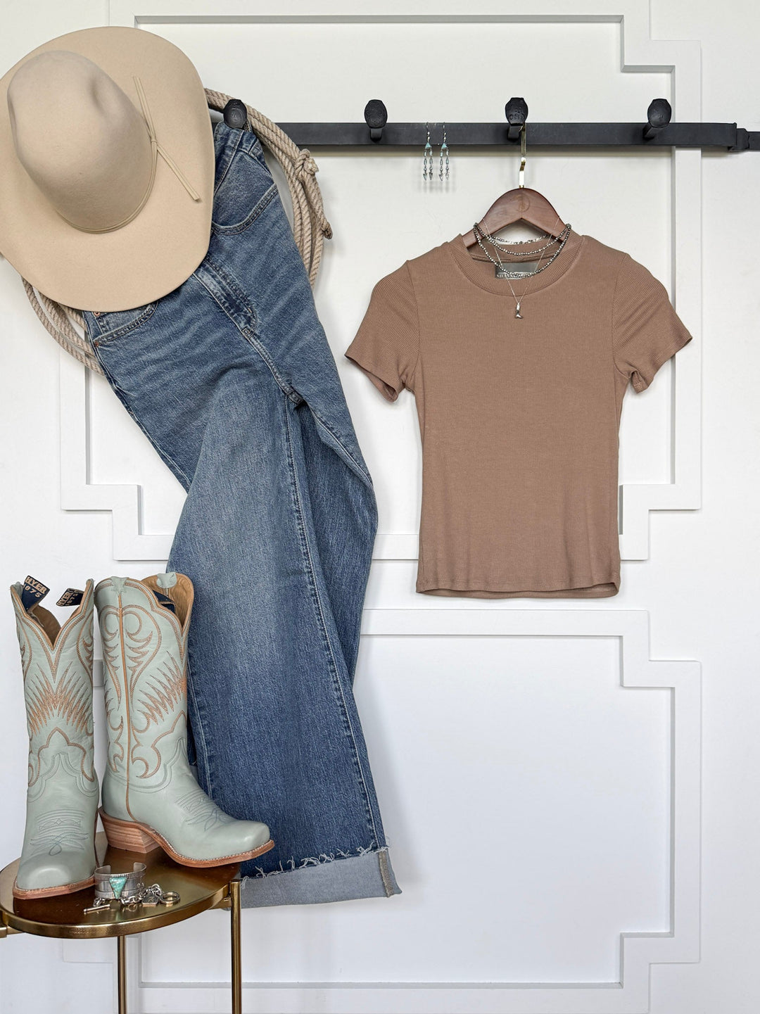 A Lamotte Beige Ribbed Tee by Savannah Sevens western life{&}style hangs on a rack next to blue jeans and a beige cowboy hat. Light blue cowboy boots are displayed below, with jewelry and accessories arranged on a small round table nearby.