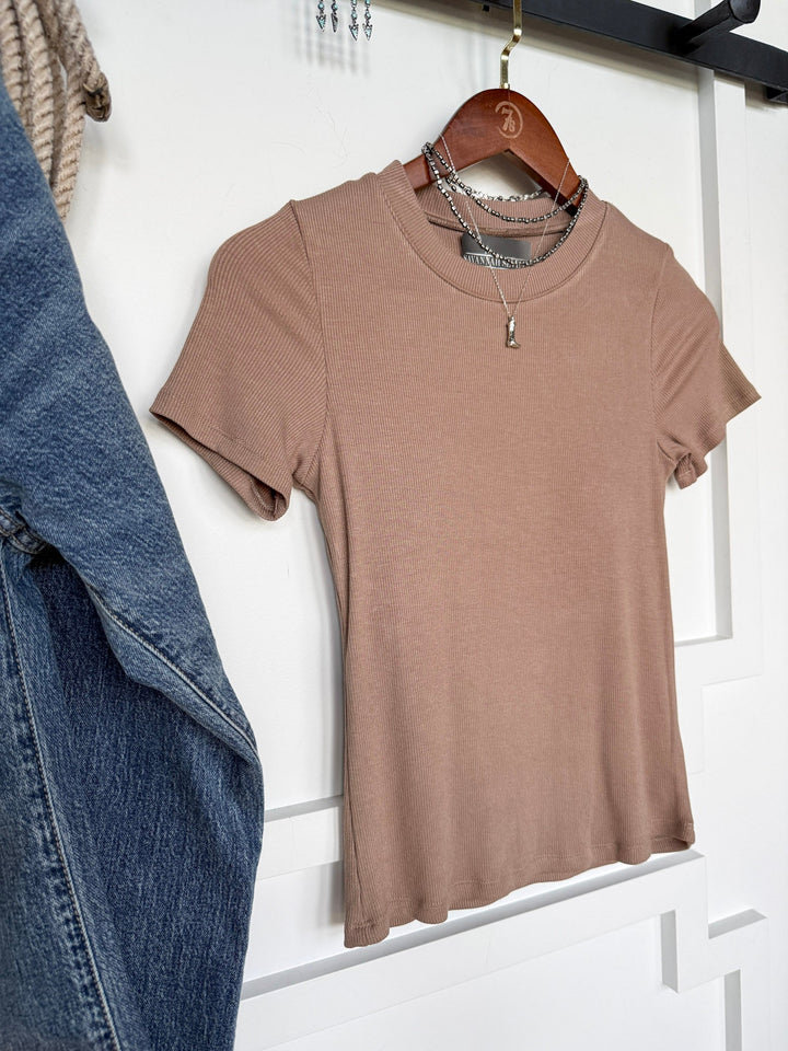 The Lamotte Beige Ribbed Tee by Savannah Sevens western life{&}style, a short-sleeve ribbed built-in bra top, hangs on a wooden hanger layered with silver chain necklaces against a white paneled wall beside blue jeans.