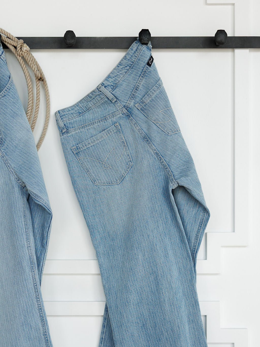 The Landry High Rise Slim Wide Leg Jean in light blue pinstripe by Savannah Sevens western life{&}style hangs on a black hook against a white paneled wall. Another pair of jeans and a coiled rope appear partially on the left.