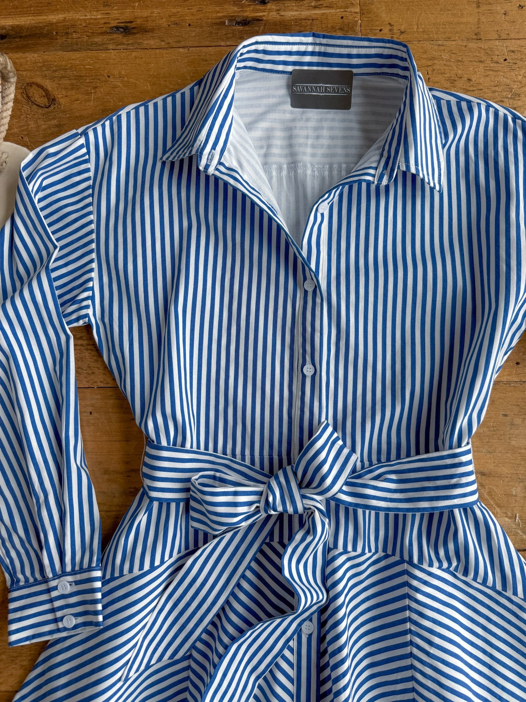 The Laneville Blue Striped Button Up Dress by Savannah Sevens western life{&}style, featuring blue and white vertical stripes, a collared neckline, long sleeves, front buttons, and a matching waist belt, is laid flat on a wooden surface.