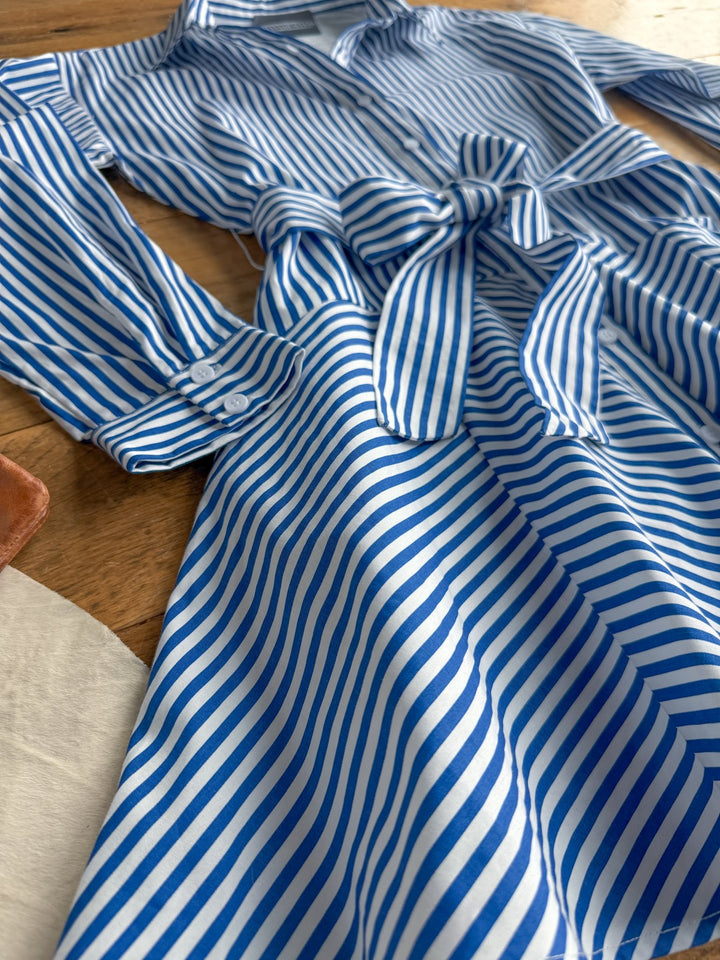 The Laneville Blue Striped Button Up Dress by Savannah Sevens western life{&}style—a blue and white vertically striped cotton dress with long sleeves, buttoned cuffs, and a matching belt—is laid flat on a wooden surface.